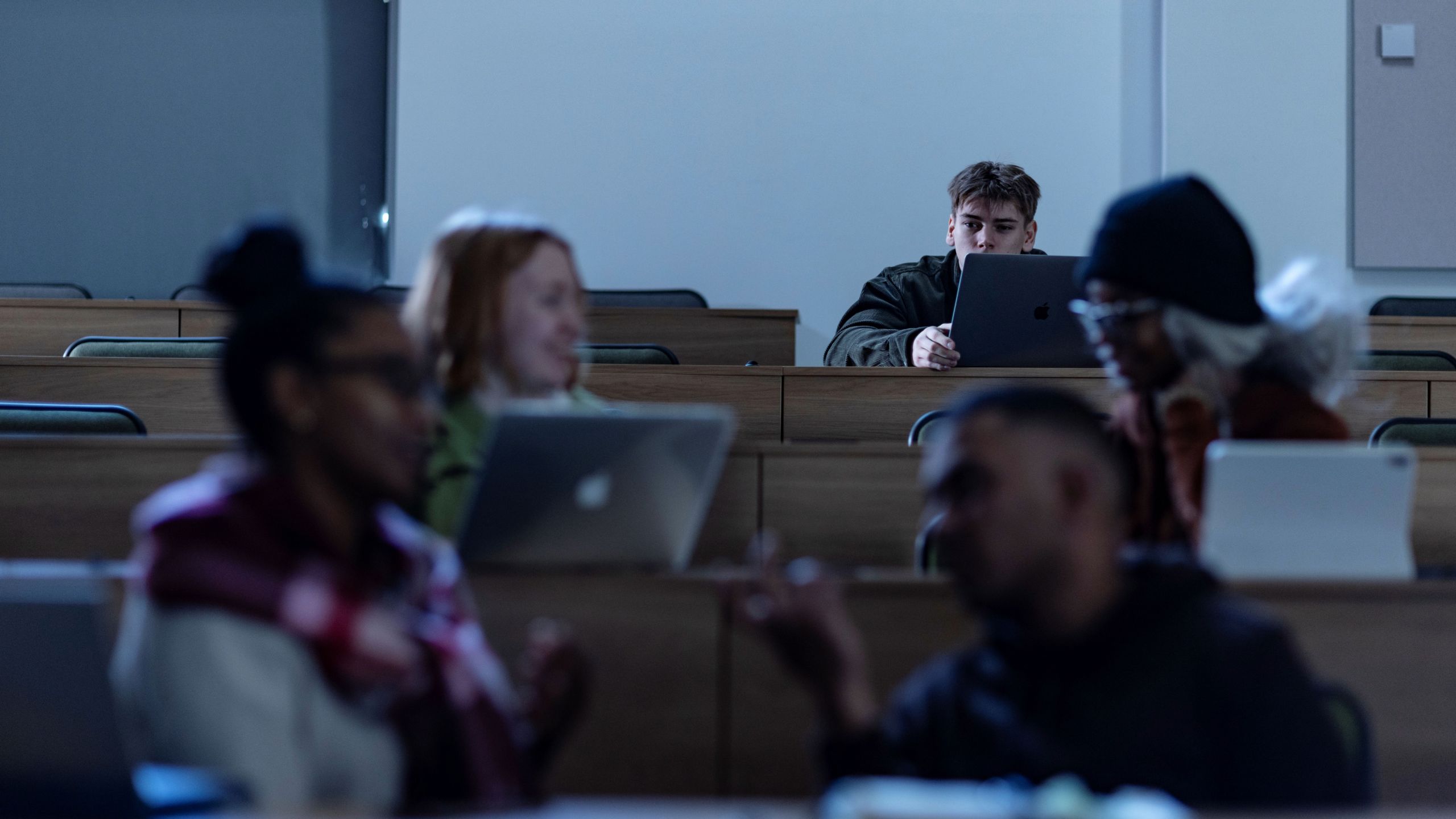 Students sit in rows of a lecture theatre, chatting with laptops. In the background, a student sits on their own, looking at the others.