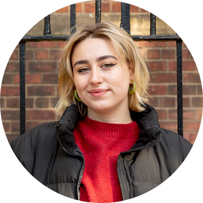Headshot of Amara. She is looking at the camera and is wearing a red jumper with a black puffer coat. She has short, blonde hair.