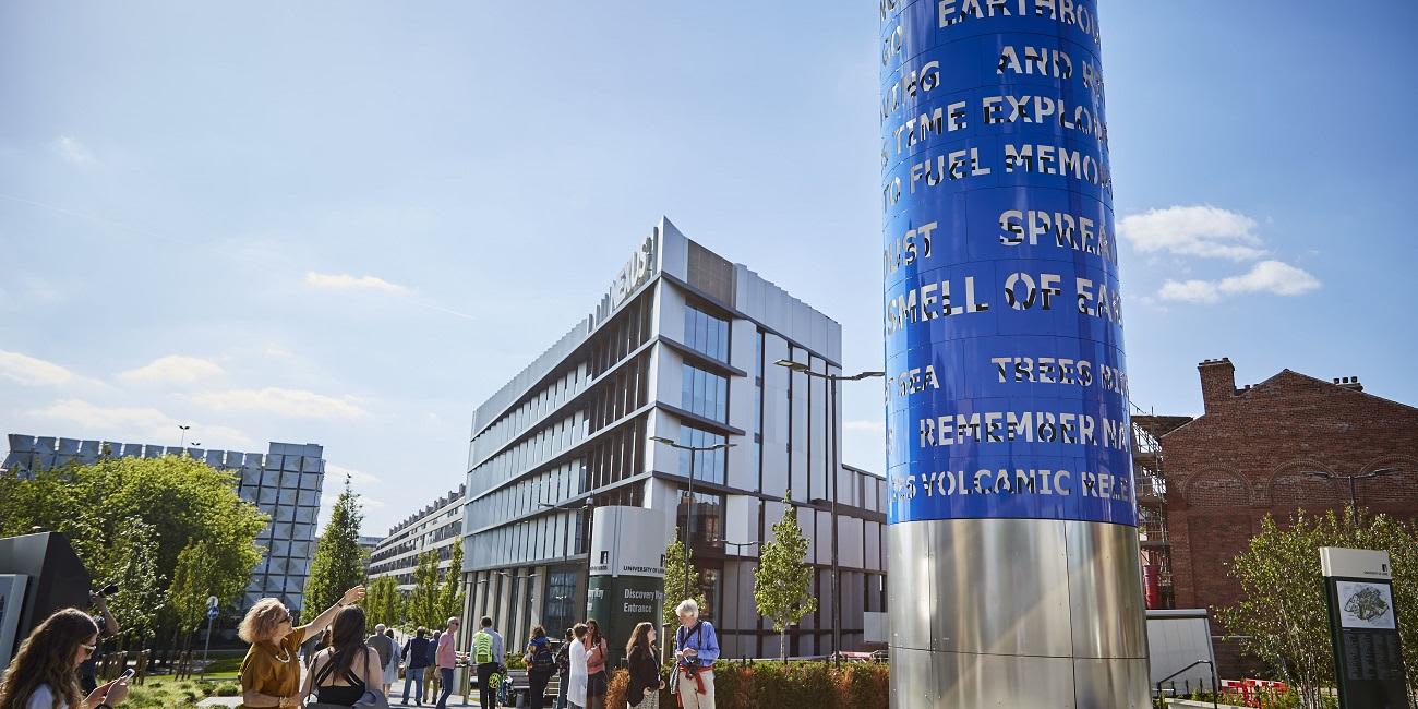 A tall blue metallic column, a sculpture on campus, stands against a blue sky. People looking up at read the words.