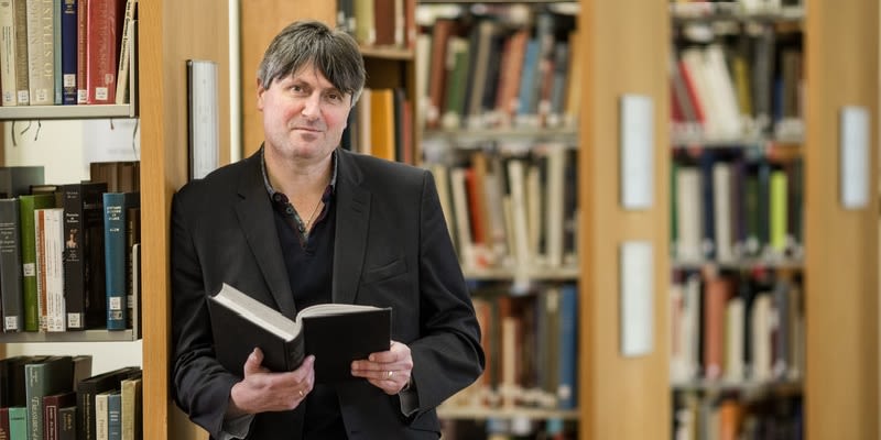Simon Armitage leans against a bookcase in a library.