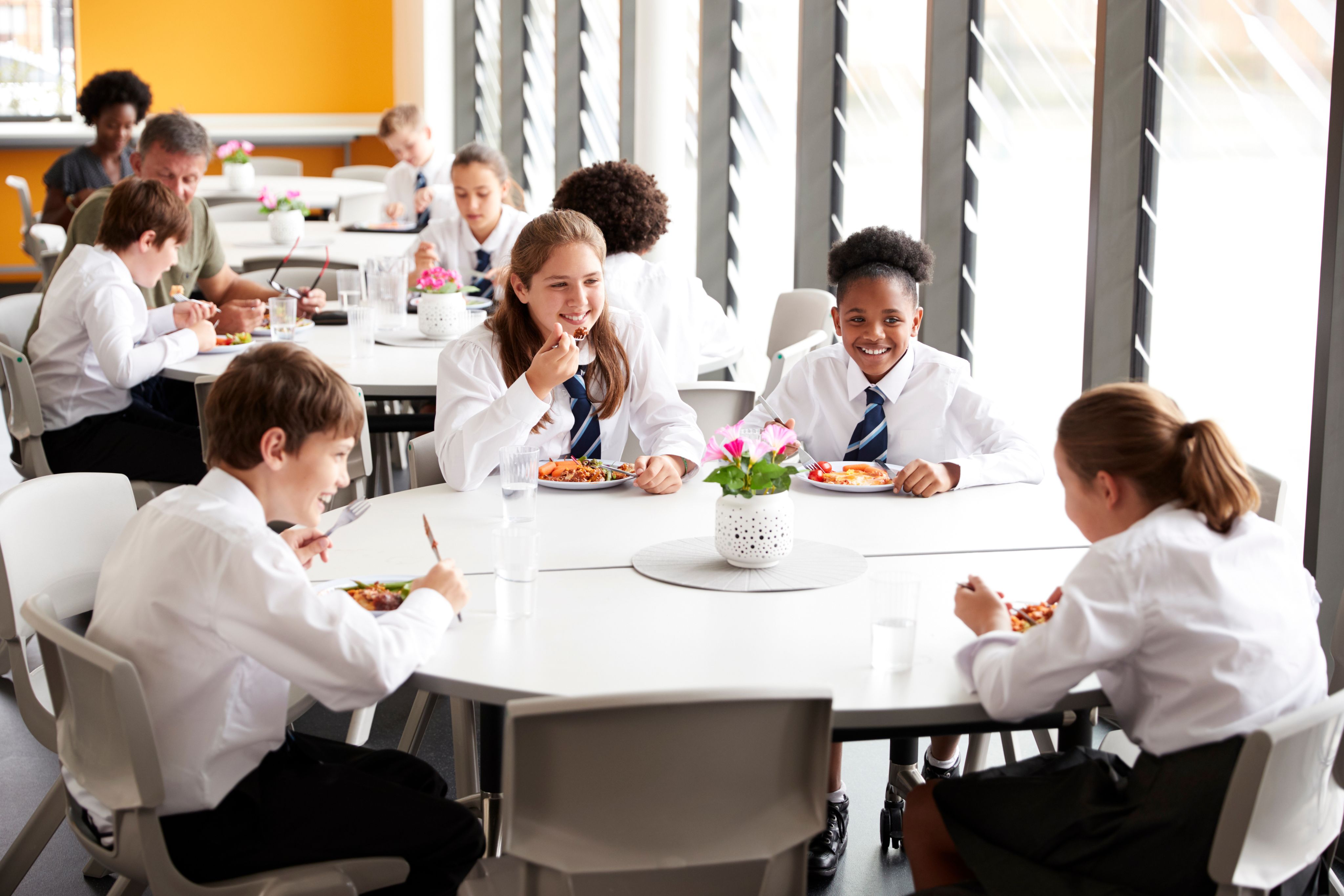 A group of secondary school pupils in uniform sitting around a table eating lunch in a canteen.