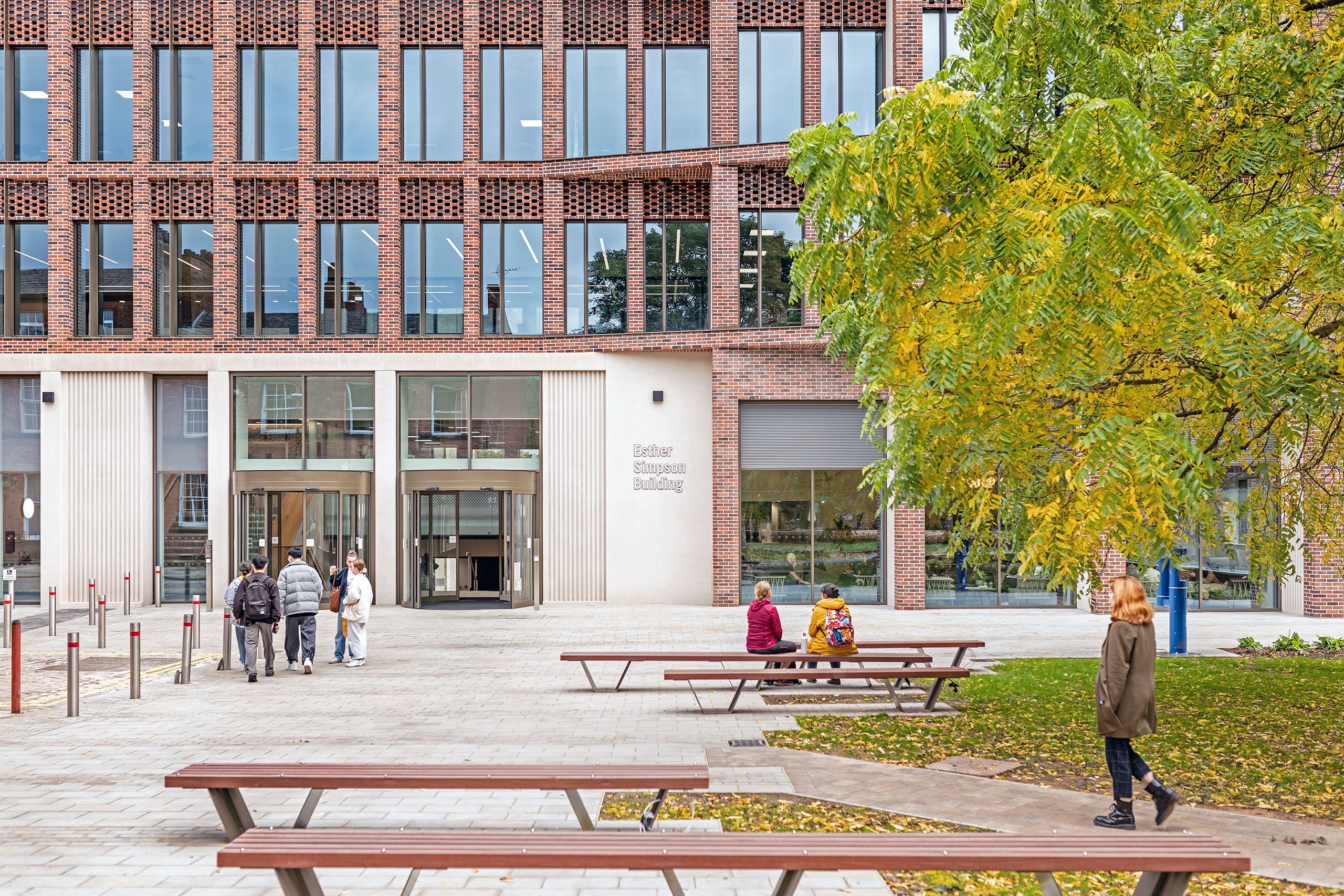 External view of the Esther Simpson Building. Students are sat on benches and stood chatting and smiling in small groups.