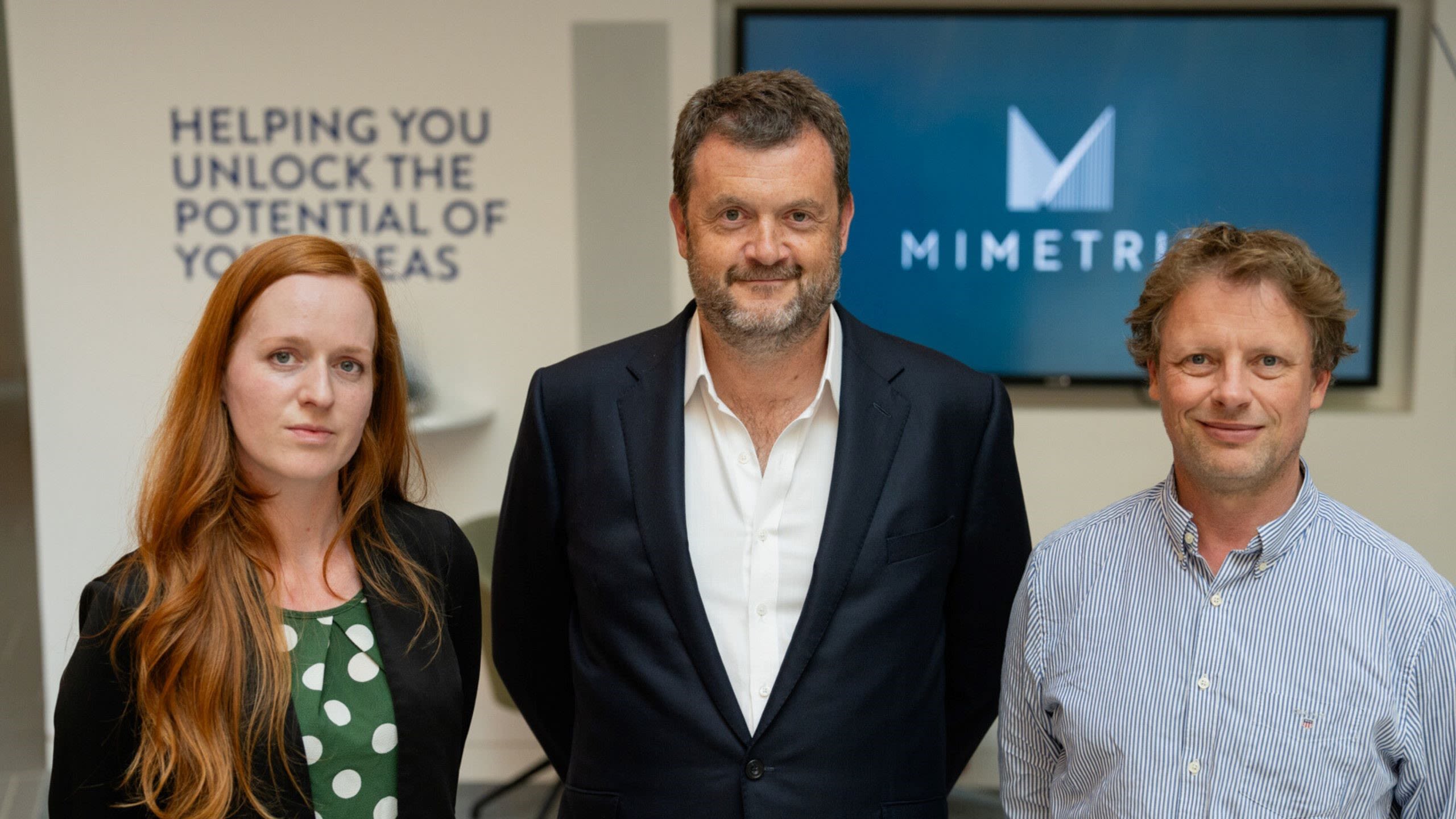 Three founders of Mimetrik stand looking at the camera, in front of the Mimetrik logo.