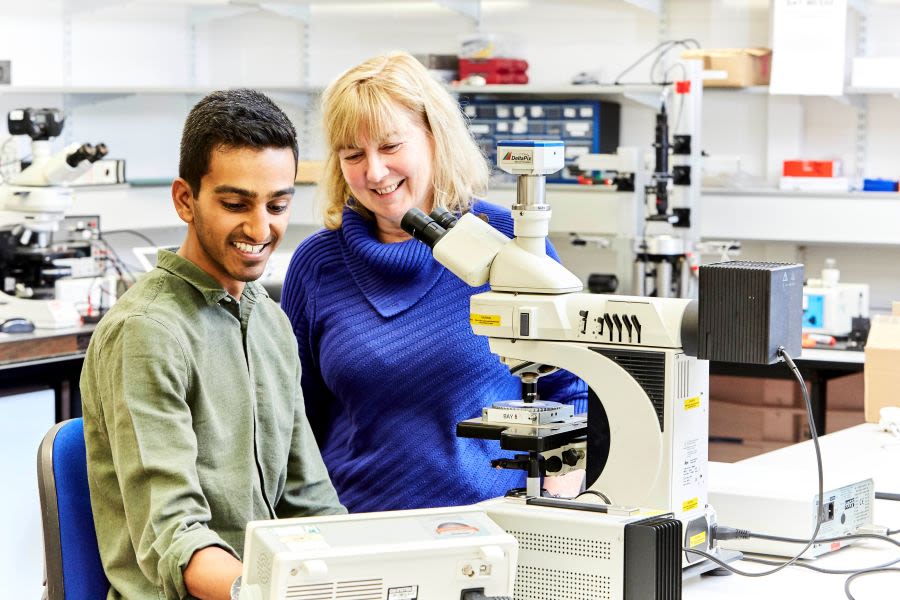 Two people stand in a lab, with a microscope in front of them, smiling at a screen