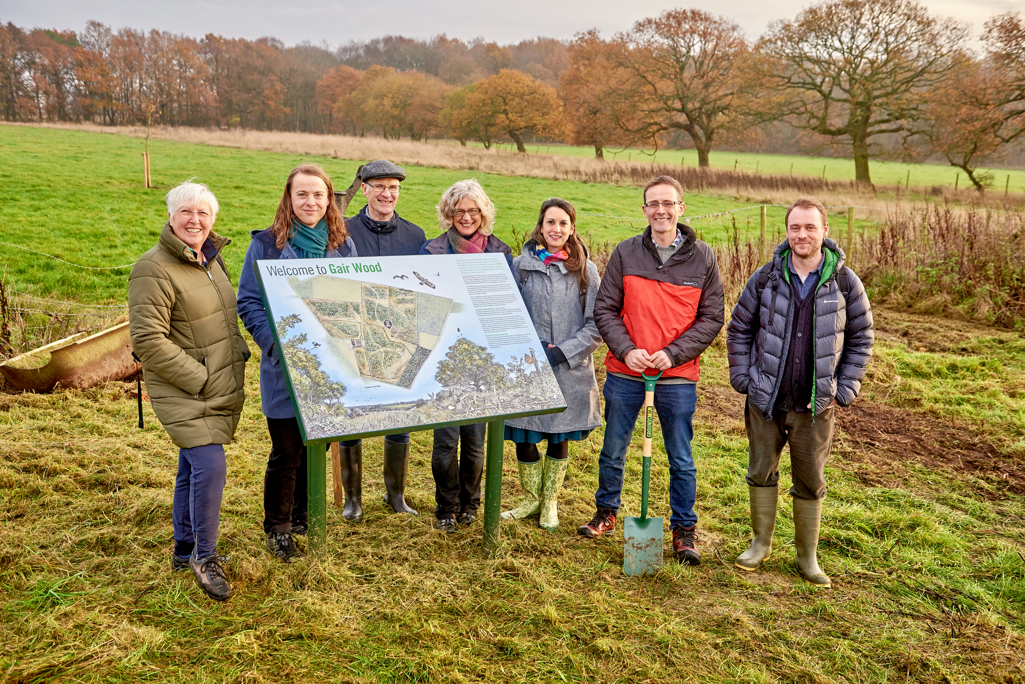 Seven people standing in a field behind an information plaque about Gair Wood. From left to right, these people are Sheila Huntridge, Robin Hayward, Steve Denison, Anna Gugan, Cat Scott, Dom Spracklen and Tom Sloan