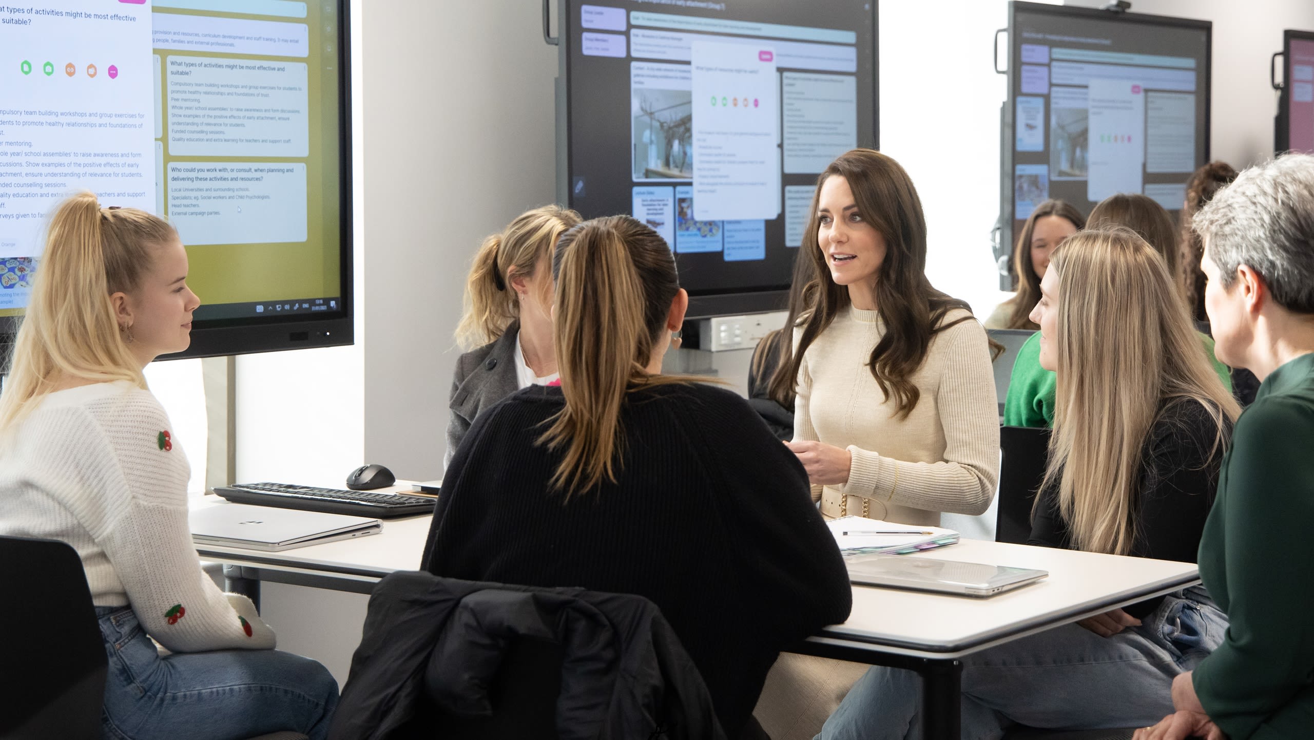 The Princess of Wales chatting to a group of people sat at desks. There are other people in the background and large screens on the wall with text and images on them.
