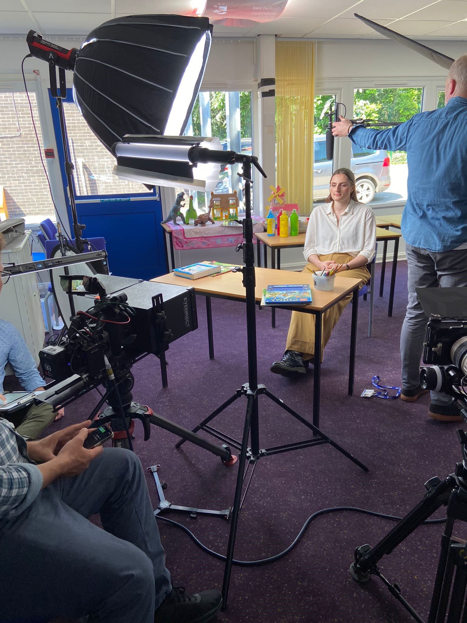 Niamh Johnson sitting at a desk in front of a large light and a camera for filming, and a technician adjusting a microphone boom.