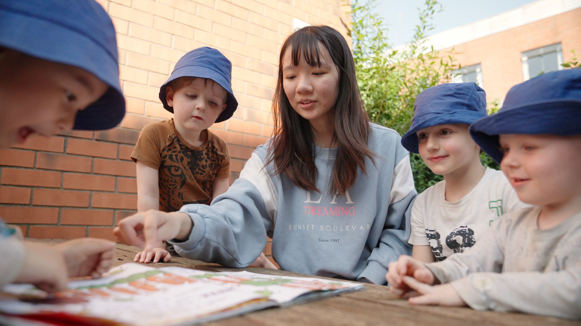 Leeds student Hongqin You interacting with four young children around a table outside, pointing at a book.