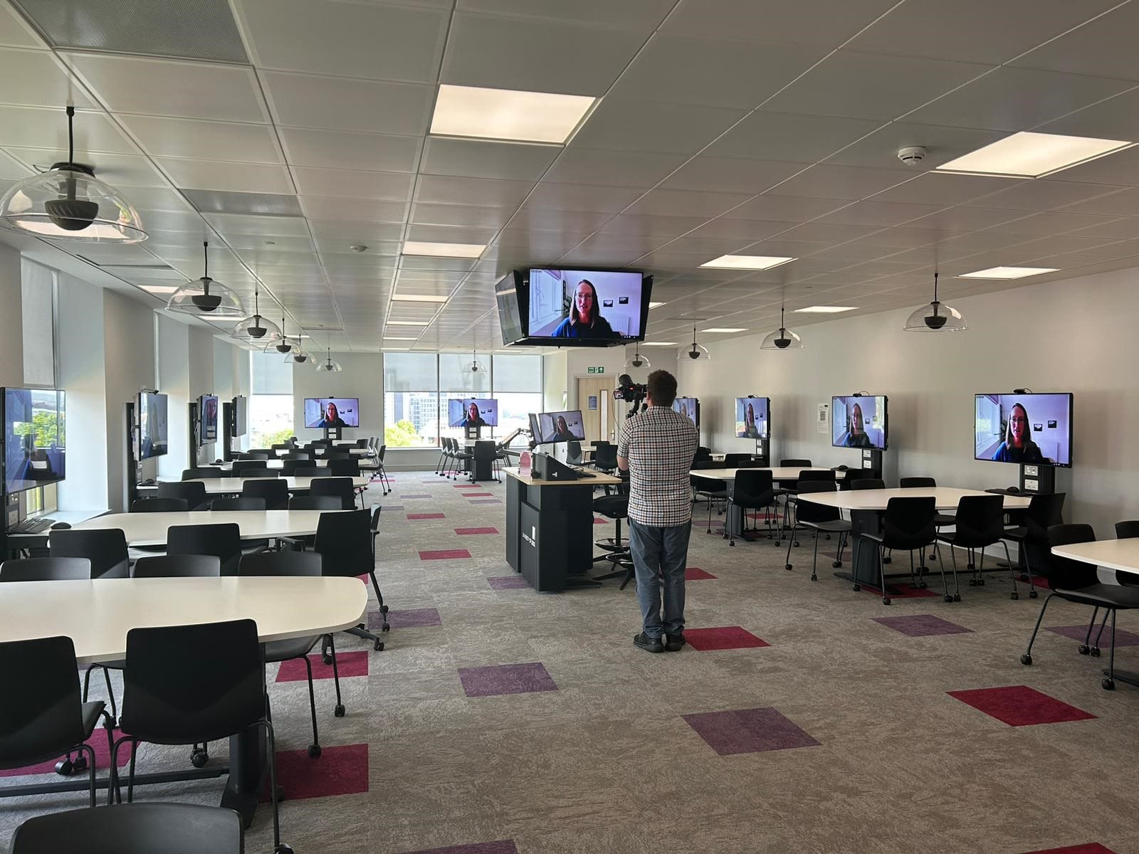 Sarah McMullen is on display screens around a teaching room. Another person stands holding a camera for filming.