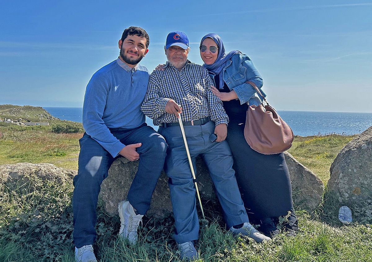 Three people sat smiling on a grassy edge, with sunshine and sea beyond.