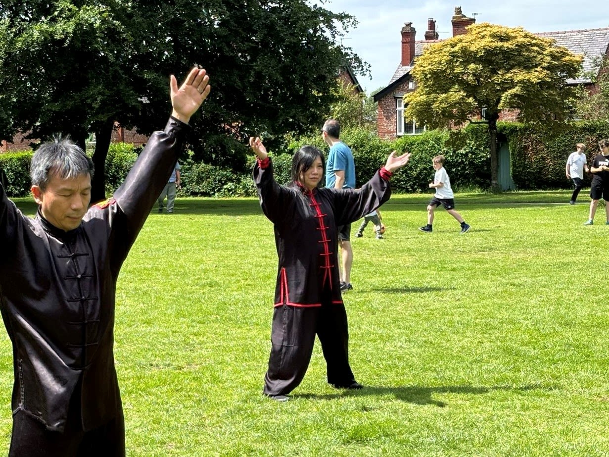 Sally demonstrating Tai chi outside on the grass. The sun is shining and children are playing in the background.