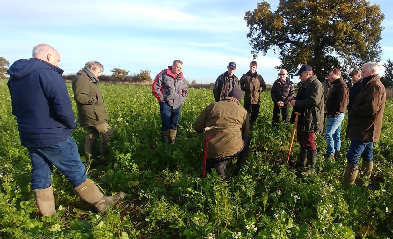 a group of farmers standing in a circle in a field of crops. They are looking at a kneeling man presenting to them.