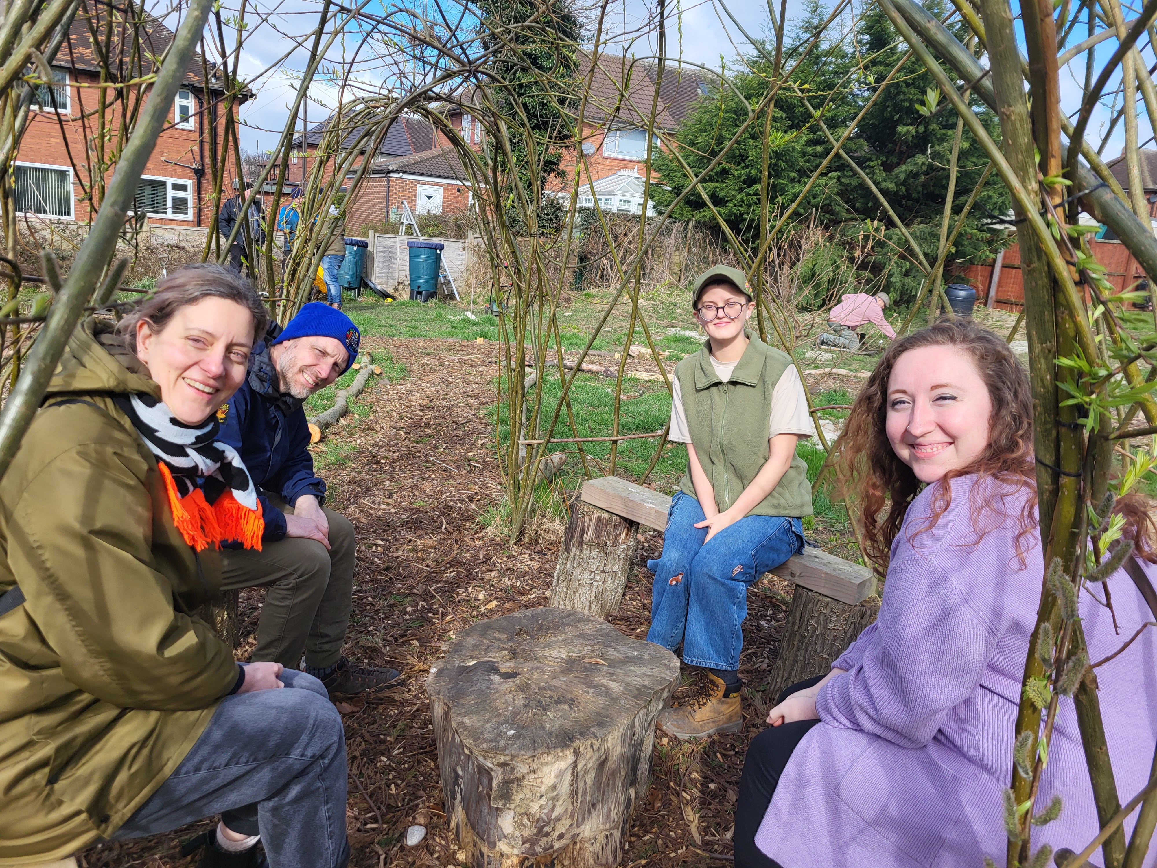 Four people sitting on benches under a structure made of willow tree stems bent into arches. The willow has no leaves so we can see the rest of the garden in the background.