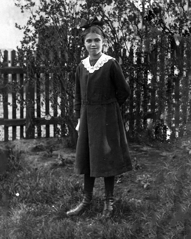 A teenage girl stood in a garden wearing a long dark dress with a white collar. There is a fence beyond.