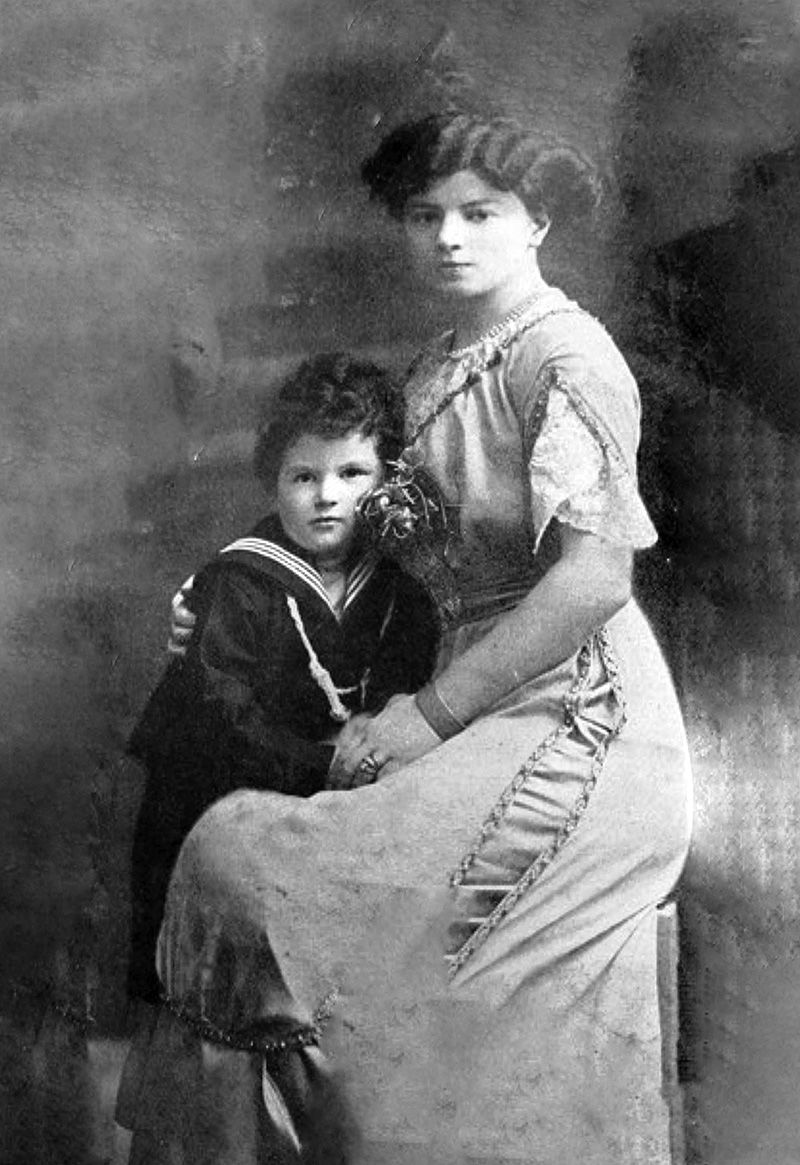 A formal, archive portrait of a woman sat on a stool in a long gown. A young boy in a sailor costume leans on her lap.