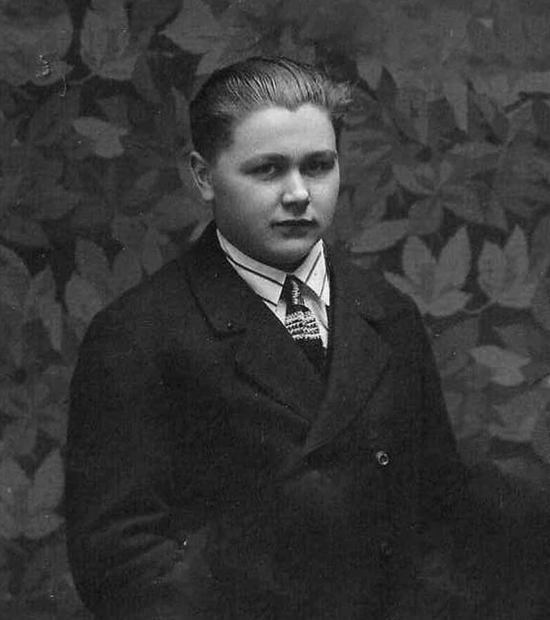 Portrait of a young man in a suit, shirt and tie in front of a leaf-patterned background.