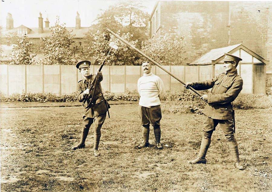 Black and white photo of two men in military uniforms facing each other with pikes with one instructor watching