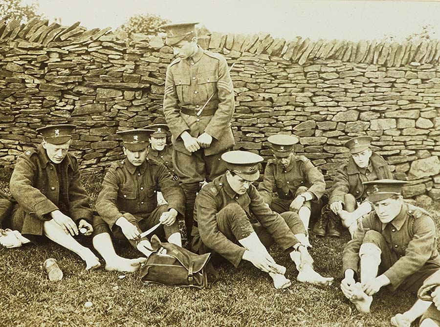 7 men in military uniforms sitting on ground inspecting their bare feet with leader standing behind them