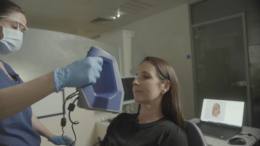 A person scanning a patient's face with machinery. The face appears on a screen behind the patient.