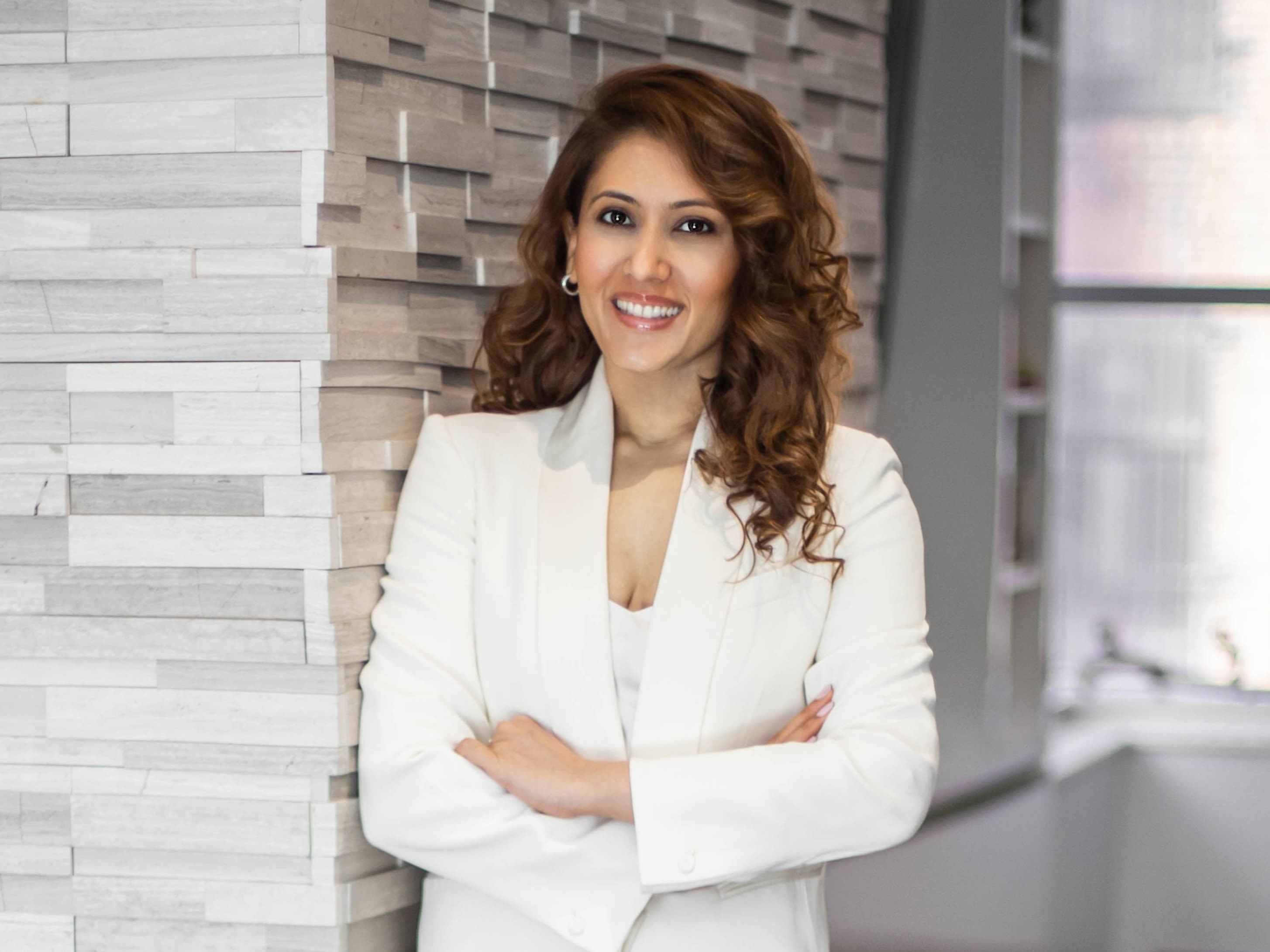 Anu Shah smiling for a photo wearing a white blazer and leaning against a grey wall
