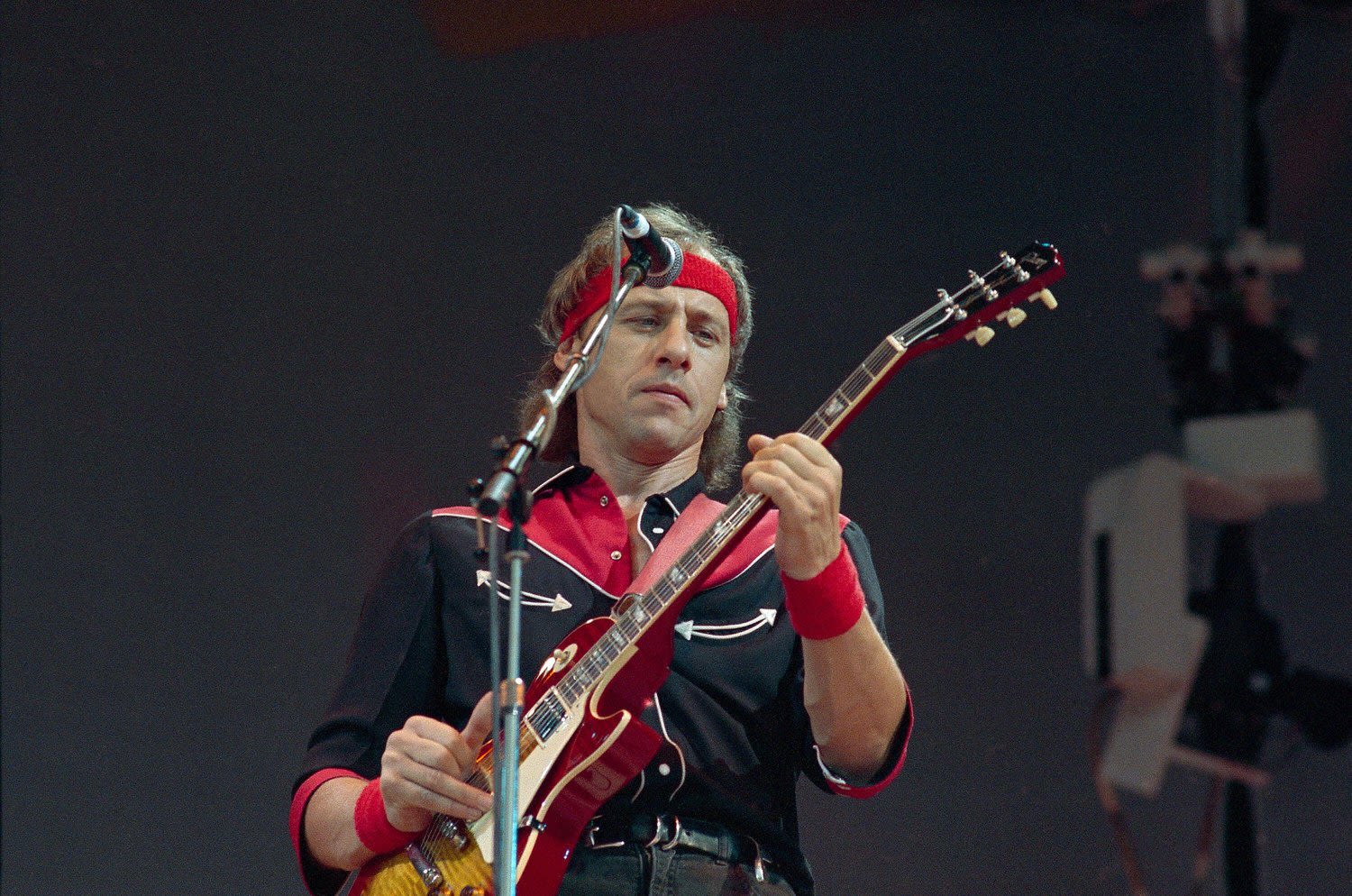 Mark Knopfler, wearing a red bandana, plays guitar on stage