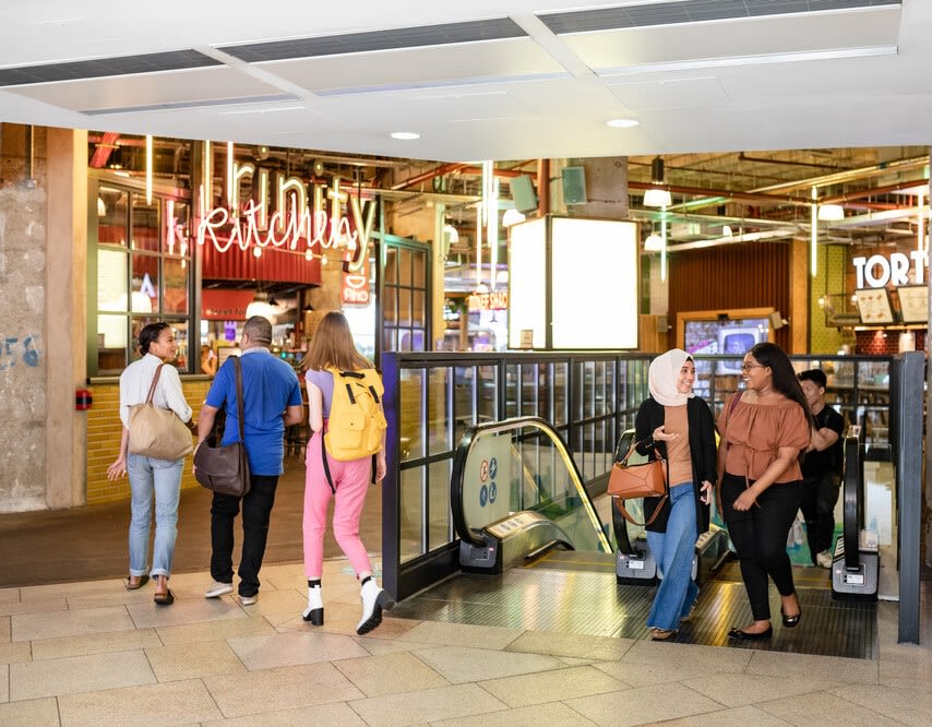 People walking in the Trinity centre. A neon sign says Trinity Kitchen