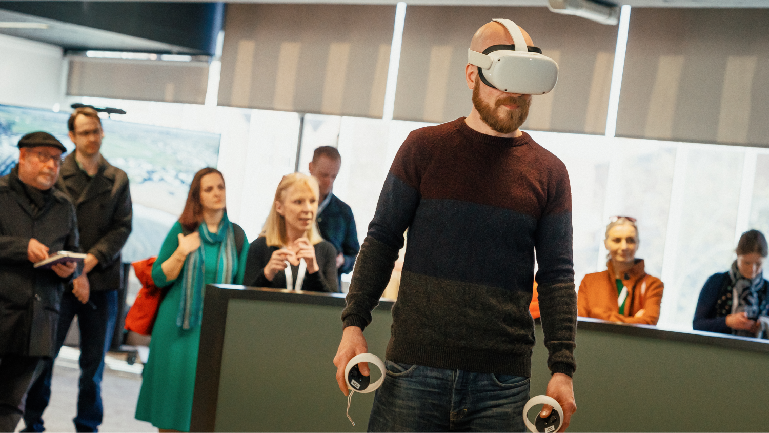 A man stands using a VR headset with a crowd of onlookers watching from behind 