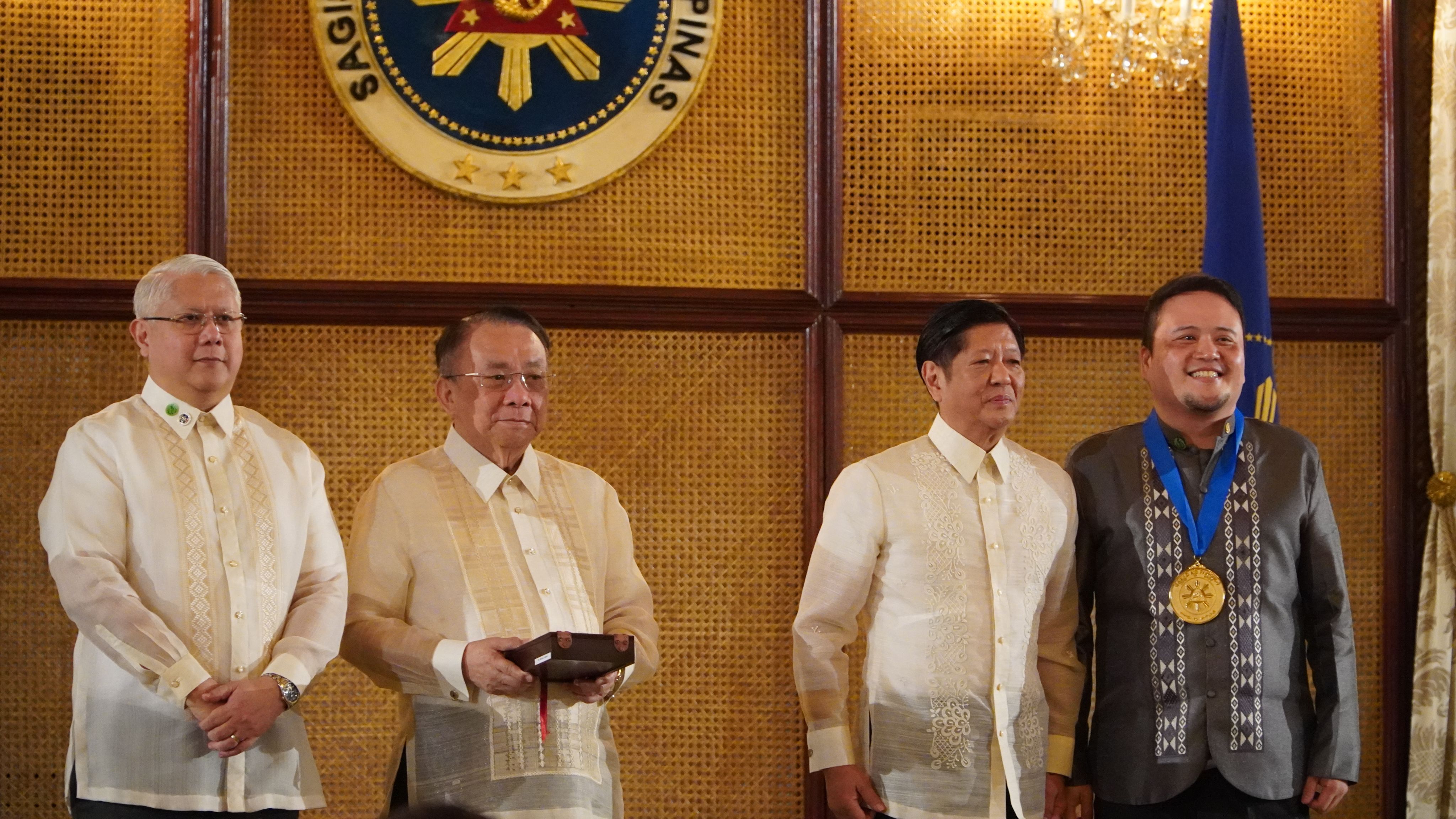 Four men stand together on stage at a ceremony, one wearing a medal