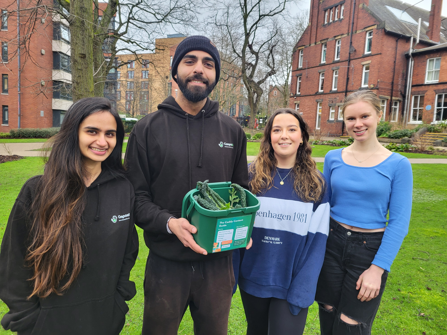 Four individuals stand side by side in a green garden and one of them is holding a box of vegetables