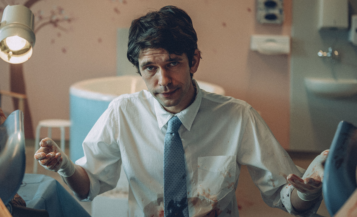 Ben Whishaw holds his arms up, wearing a shirt that is splattered with blood