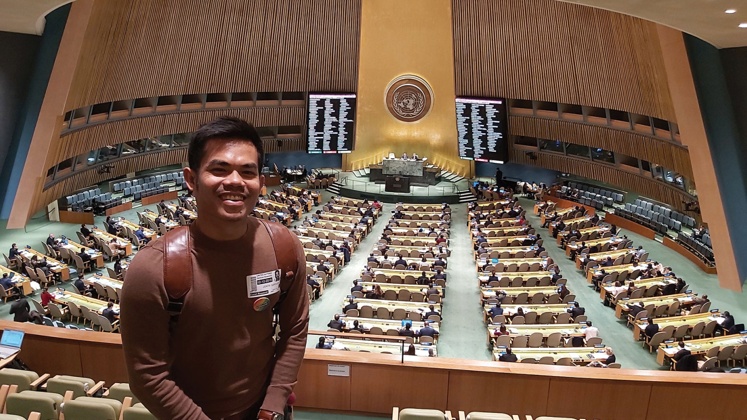 Jonta stands at the back of a conference room which is lined with desks leading to a central stage