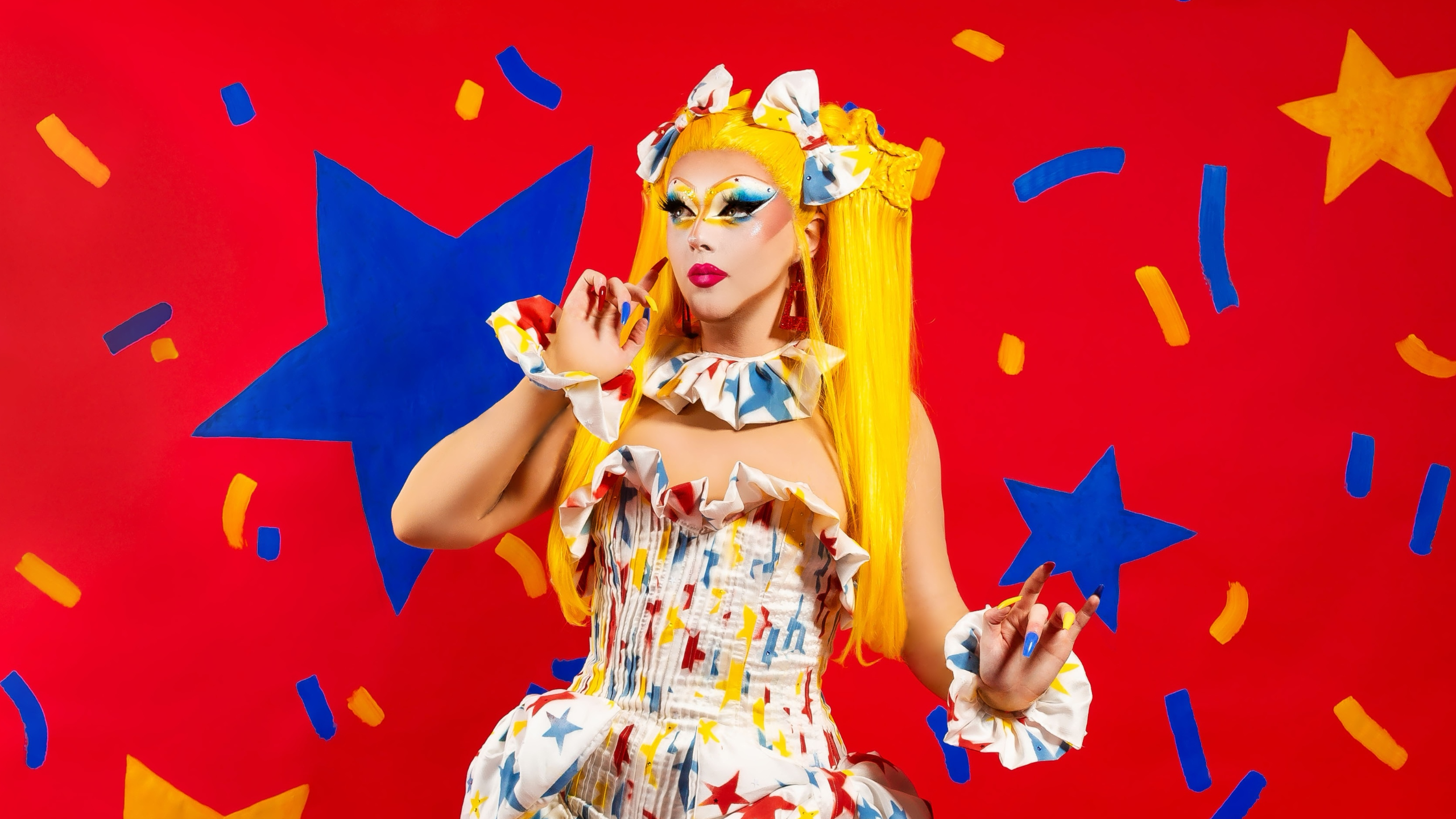 Charra Tea stands in a vibrant dress with long blonde hair. She stands in front of a red display with blue stars