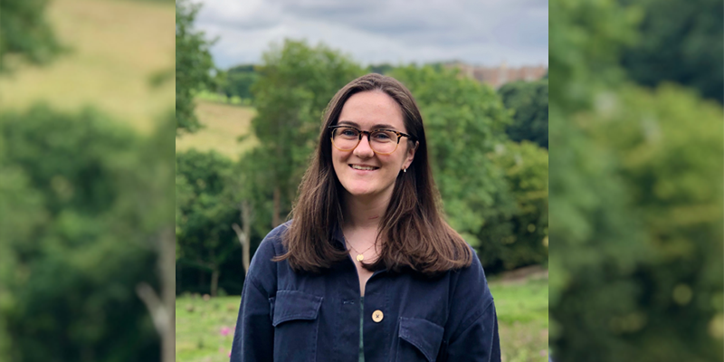 Lydia Hutton, music student, smiling. Grass and trees are in the background.