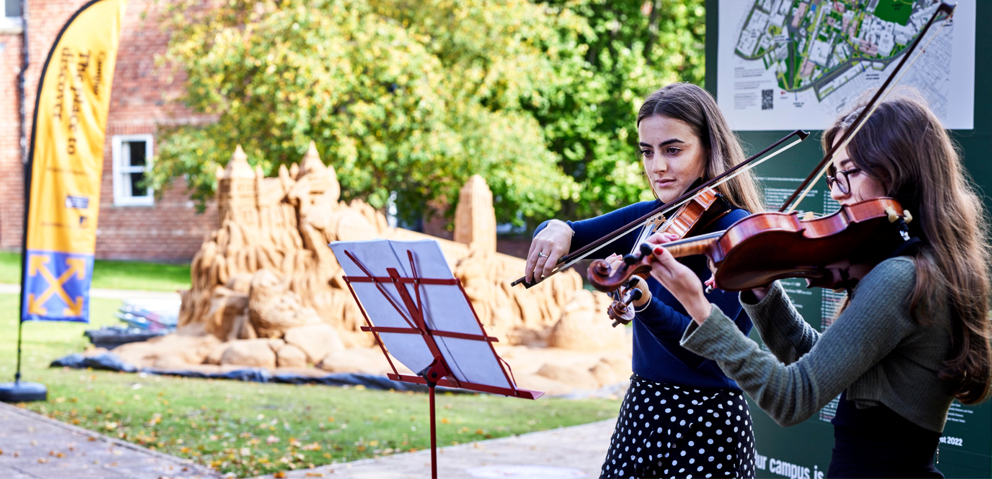 Two violinists perform on the grass before the sand sculpture