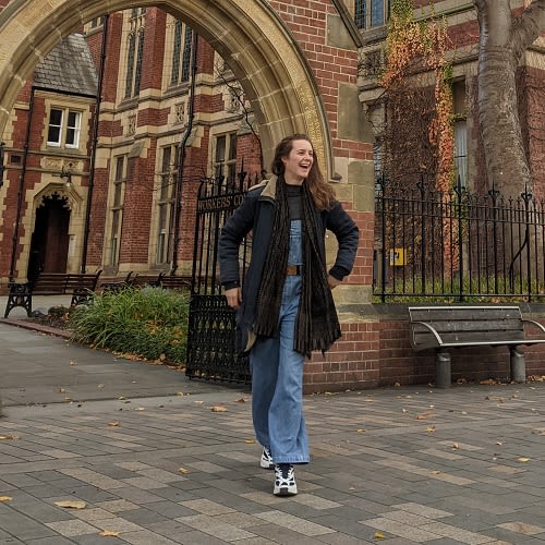A woman in denim dungarees and a black coat photographed outside the great hall mid-laughing