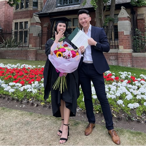 Photo of a couple taken on campus. The woman on the left is wearing a graduation gown and cap, holding a bouquet of flowers. The man on the right is wearing a suit and holding a graduation certificate.