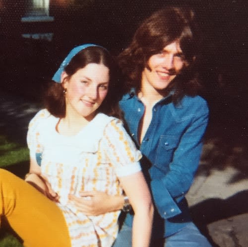 An old photo from the 1970s showing a man and woman sitting on a wall. Both have long brown hair. The man is wearing a denim shirt and the woman a white and orange blouse.