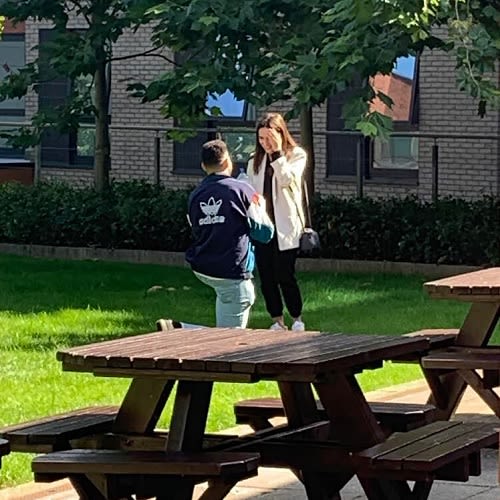 A photo of a man on one knee proposing to a woman. He wears an adidas jacket and she is wearing a white coat.