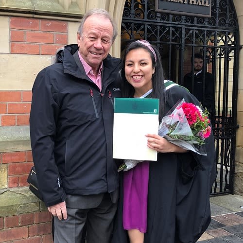 A man and a woman pose for a photo outside the great hall. The woman on the right is wearing her graduation gown and holding flowers and her certificate