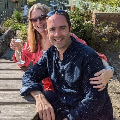 A couple sat in a beer garden. The woman sits behind the man with her arms around him. She is holding a champagne glass