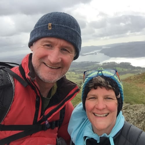 A selfie of a couple hiking. Both smile at the camera. Both are wearing wooly hats and walking gear.