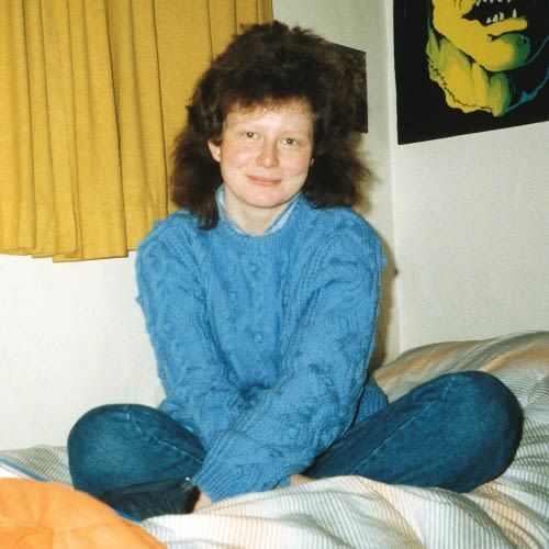 An old photo from the 1980s. A woman with long dark hair sits crossed legged on a bed. She is wearing blue jeans and light blue jumper.