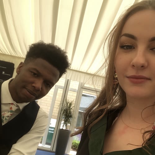 A selfie photo of a couple. The woman is in the foreground and the man is in the background. Both are looking at the camera.