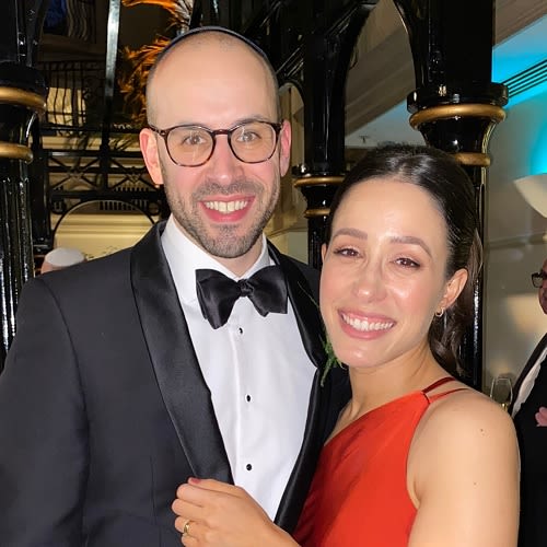 A couple posing a smiling for the camera. The man on the right wears glasses and a tuxedo, the woman on the left wears a red dress
