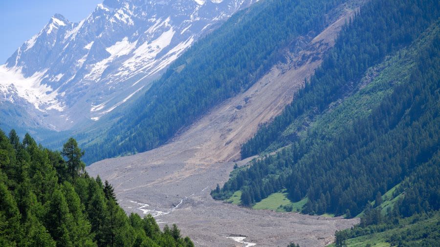 A mountainous valley with trees in the foreground and a large landslide sweeping through the valley, leaving a clearing of trees. In the background are snow-capped mountains.