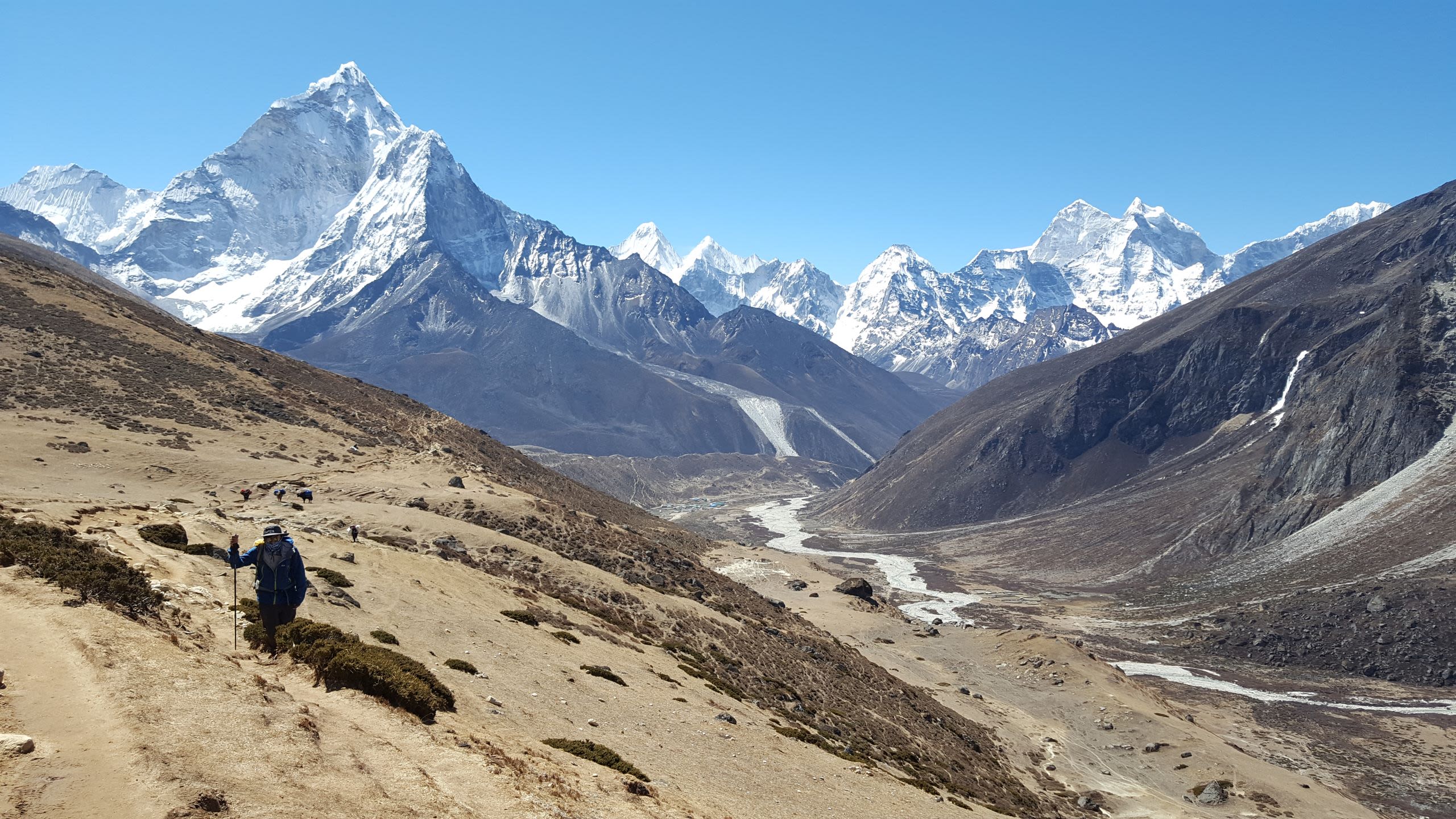 A person treks up the surface of a rocky mountain, with snow-capped rugged mountains in the background and a clear blue sky.