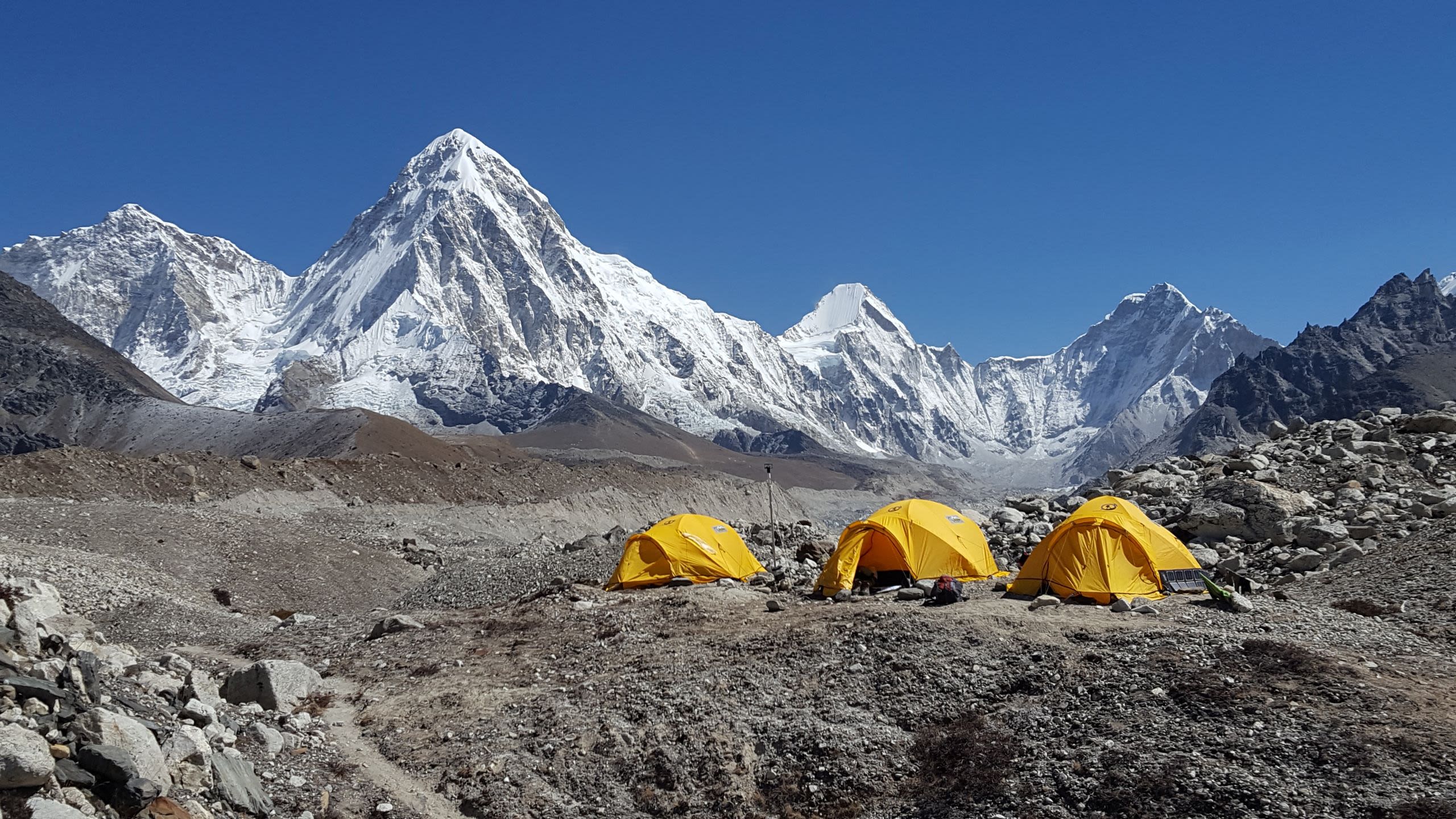 Three orange tents pitched on rocky ground with a snowy mountain range in the background, against a bright blue sky.