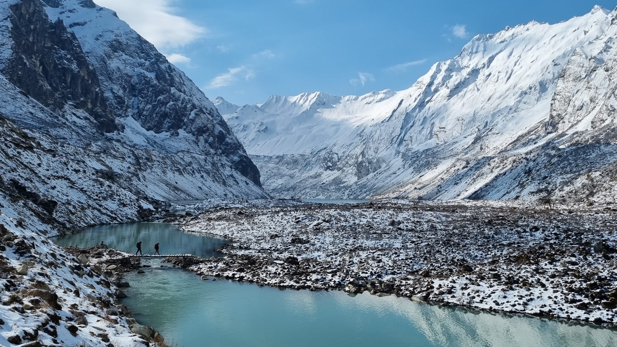 An ice blue lake meanders in a valley surrounded by snow-covered mountains. A small bridge crosses the lake, with two silhouettes of people walking across. It's a bright day with a clear blue sky.