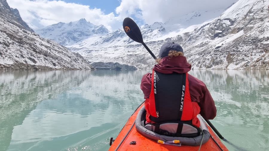View from behind of someone kayaking across a clear blue lake surrounded by snow-covered mountains.