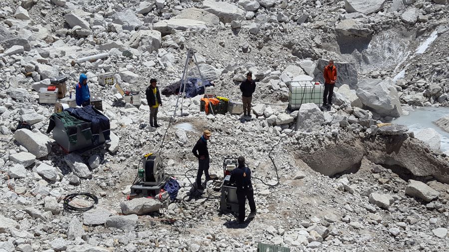 Overhead view of people standing on a rocky lapdscape with scientific equipment, including a wheel of cable, a tripod, and machinery.